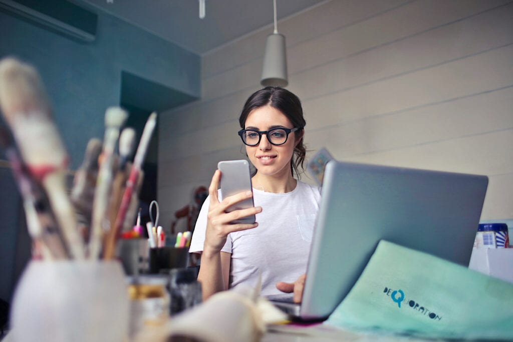 Woman sitting at her computer working with her phone with paintbrushes in the foreground