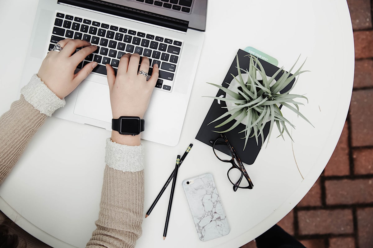 Photo of someone typing on a laptop keyboard with a plant, glasses and pencils next to the computer.