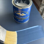 A person brushing on a coat of water-based topcoat onto a painted blue surface with a yellow bristled brush.