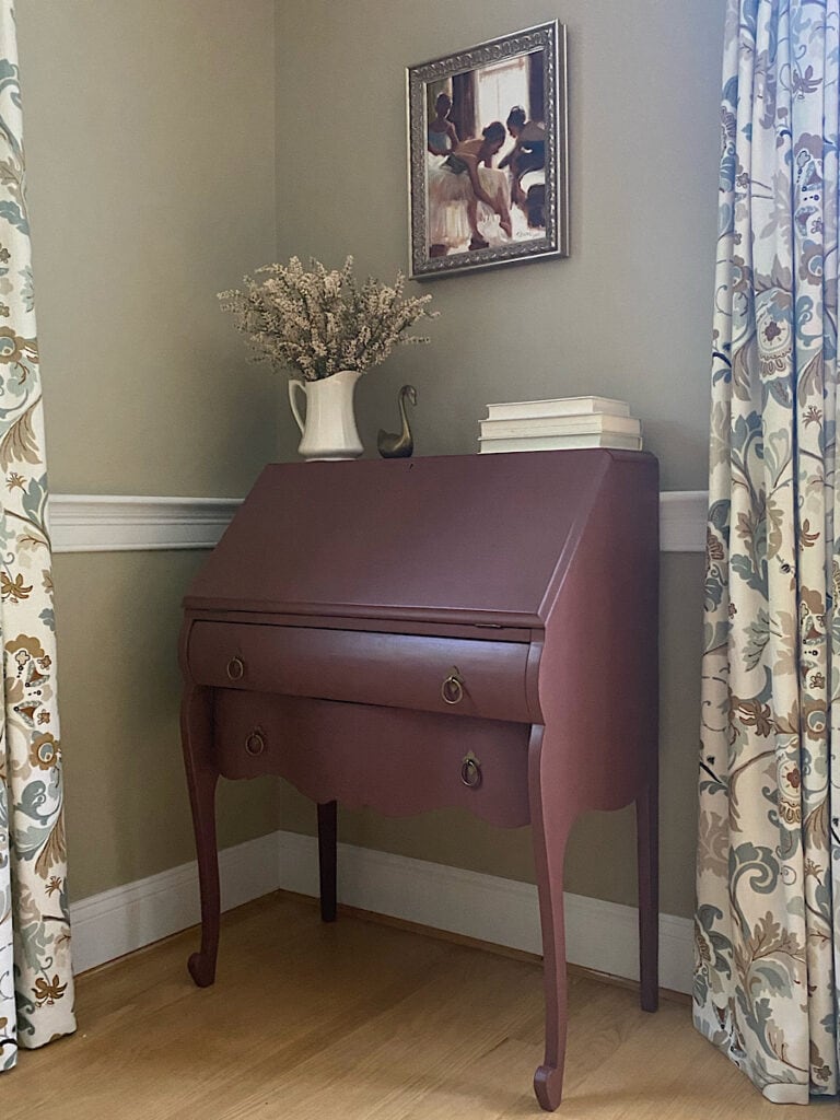 Photo of a dark red secretary desk with a tan wall, light floors and flowered drapes.