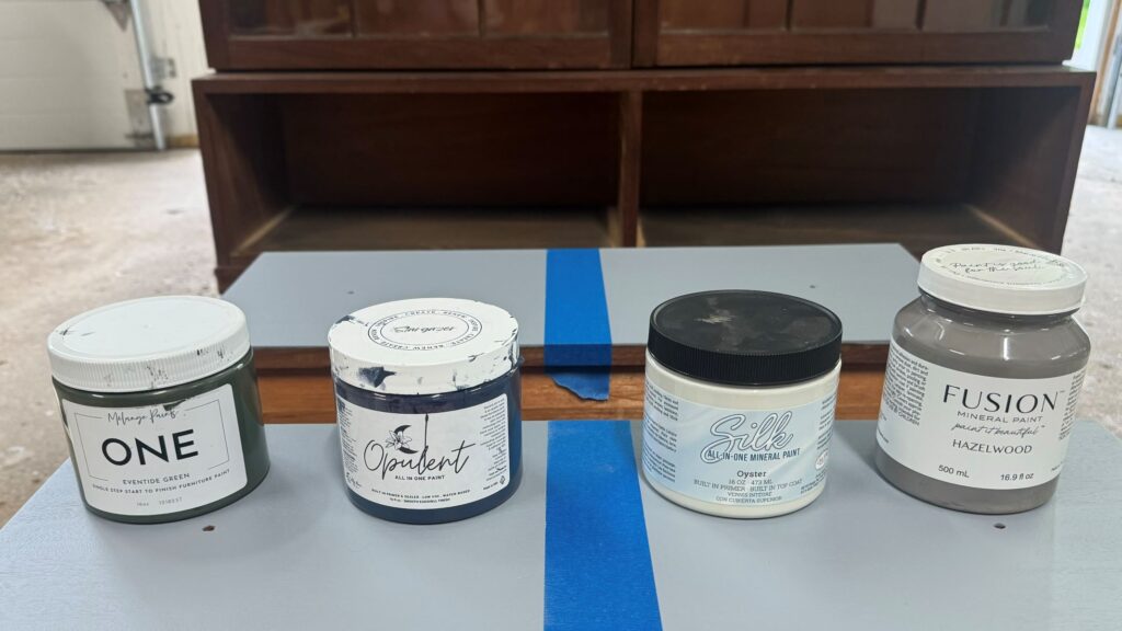 Four all-in-one furniture paints lined up on top of a primed drawer. From left to right, Melange ONE, Lilly Moon Opulent, Dixie Belle Silk, Fusion Mineral Paint.