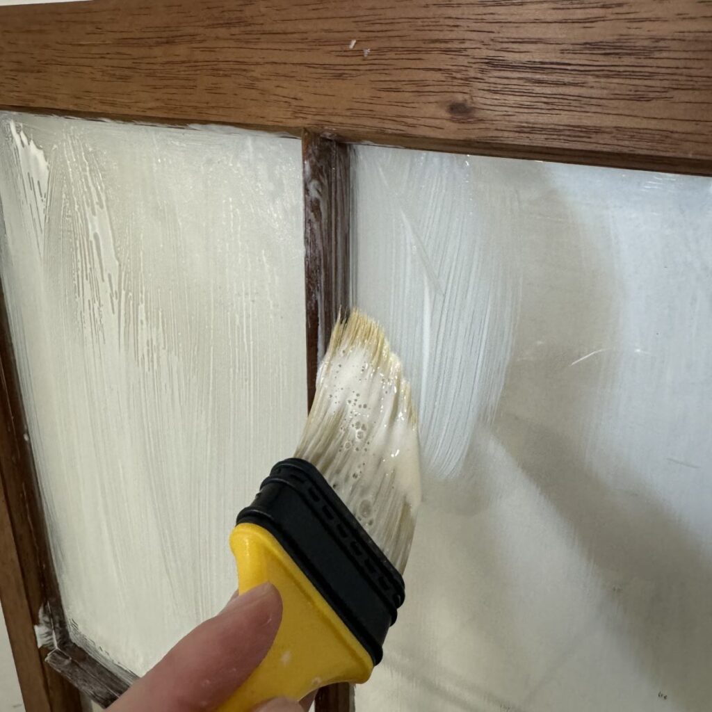 Photo of a person applying liquid mask over glass panes and wood on a cabinet door using a paintbrush.