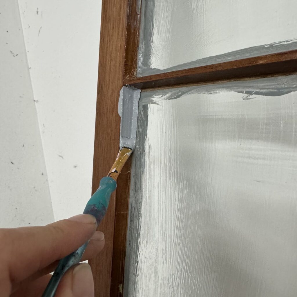Photo of a person applying paint over glass panes and wood on a cabinet door using a paintbrush. Panes and wood have been coated with a masking liquid and the paint is being painted on messy.
