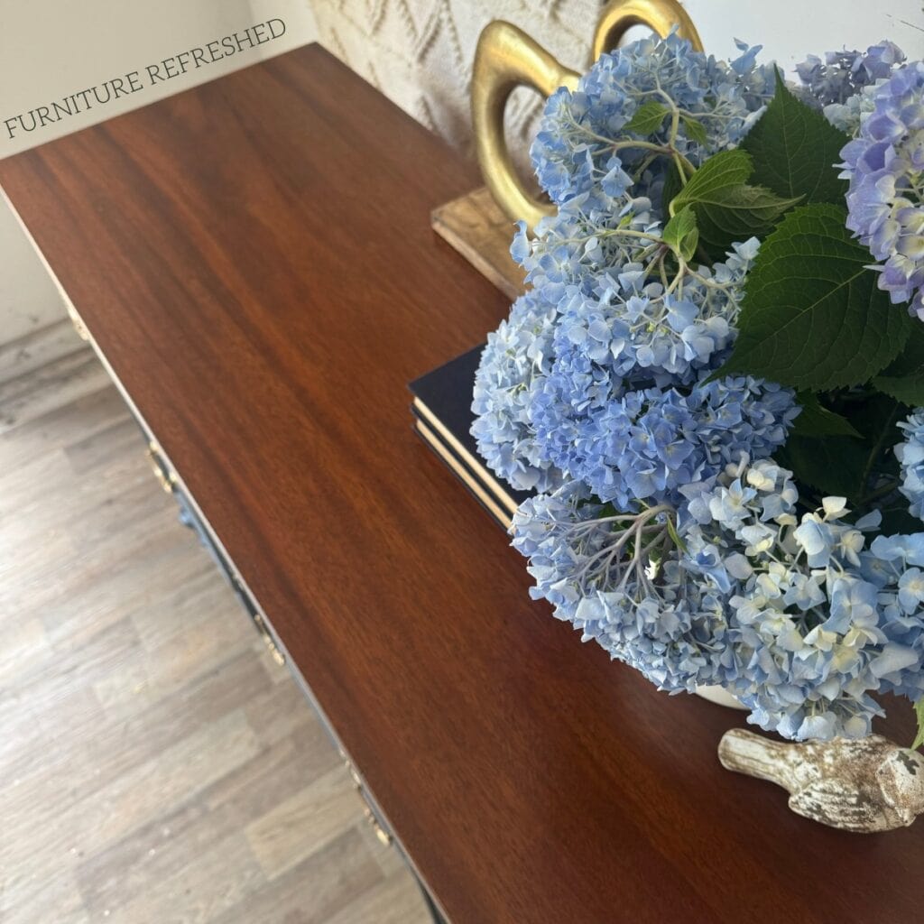 A close up of the dark wood top of a refinished sideboard. There are flowers placed on the top.