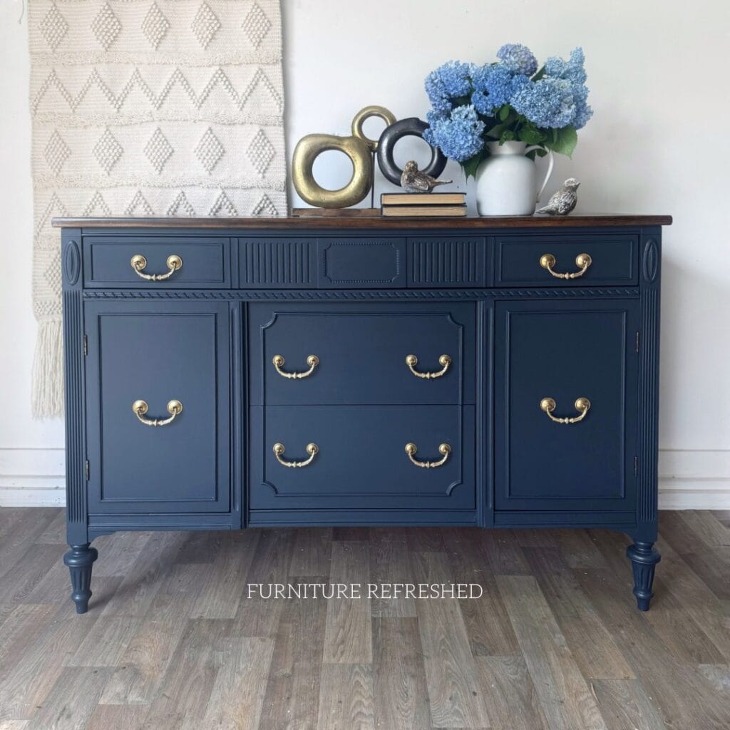 Photo of a painted navy sideboard with flowers books and other decor on top. The sideboard has brass hardware and a dark wood top.