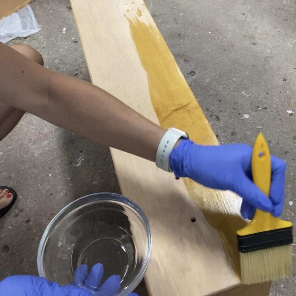Image of a vintage dresser drawer standing upright with all of the stain removed. A person with gloves is applying wood bleach to the drawer with a chip brush.