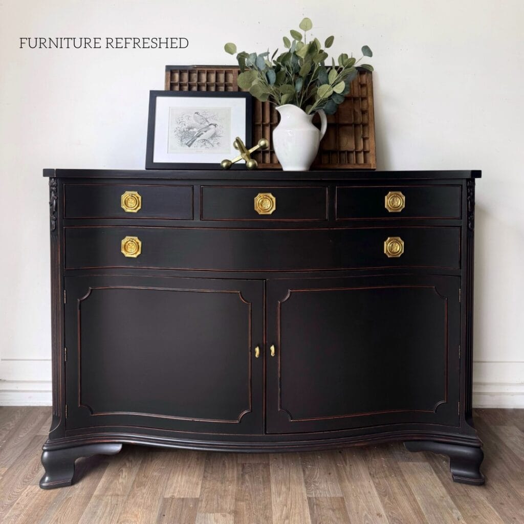 Photo of black painted sideboard with distressed edges. The sideboard has brass Hepplewhite style hardware and is pictured with a vase, artwork with birds and an old print tray resting on top.