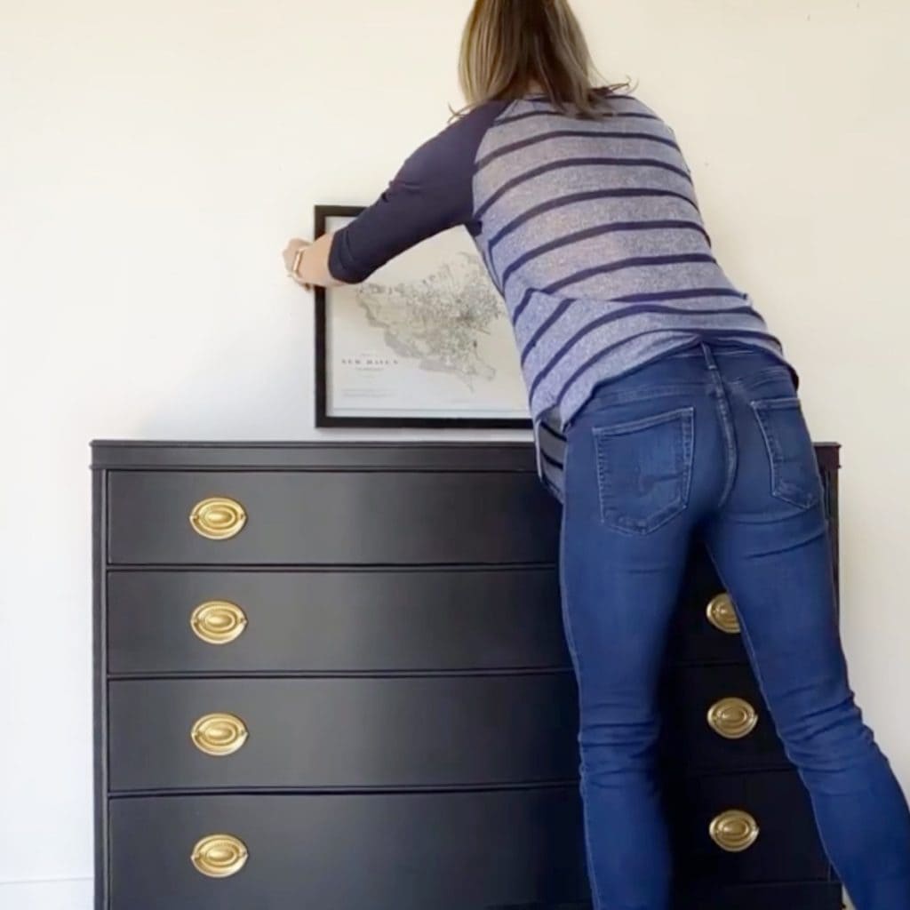 Staging a dresser. Photo of woman hanging a print over a black dresser.