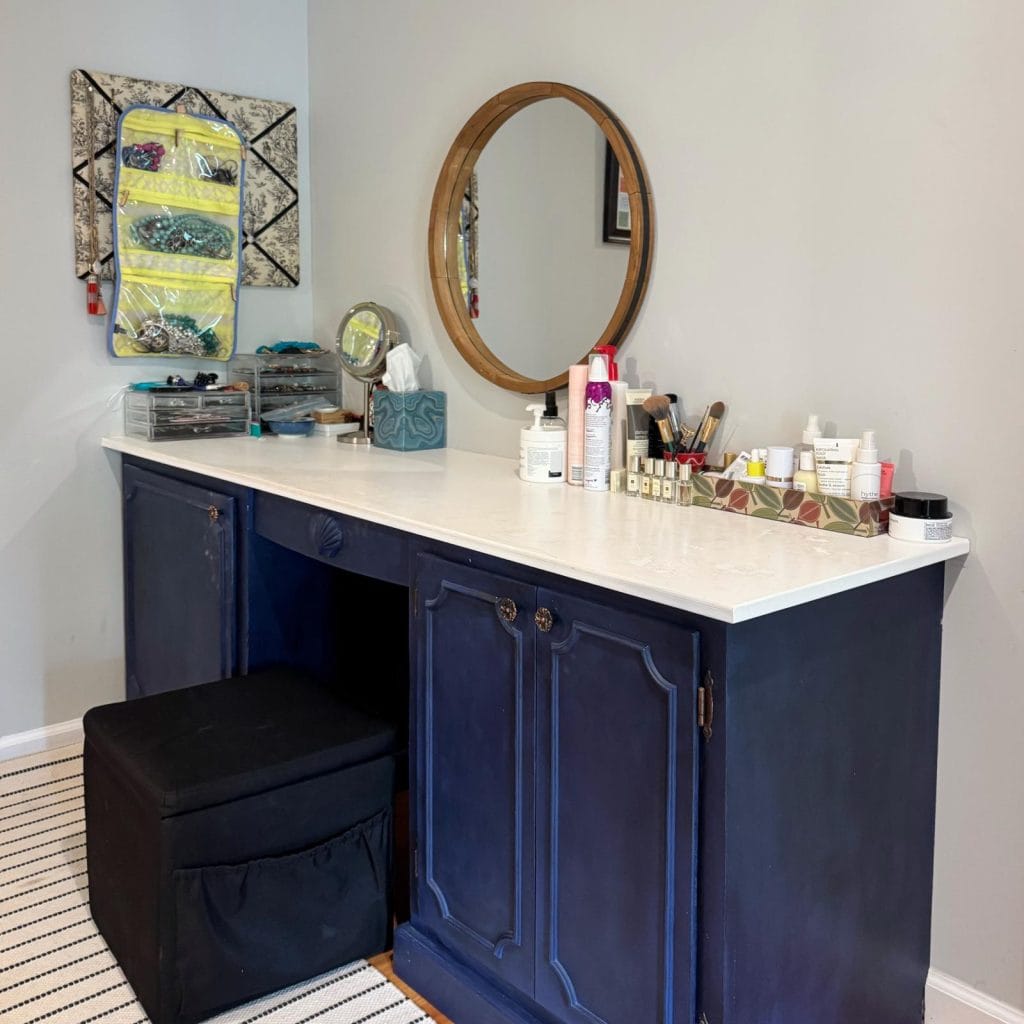 Before photo of entire vanity that is being updated. It has a painted white top and a blue bottom. There are hair and makeup products sitting on top and a circular mirror above the vanity.